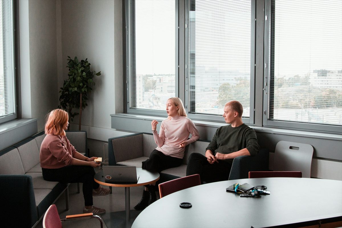 Three people having a discussion in a modern office with large windows, a round table, and a laptop.