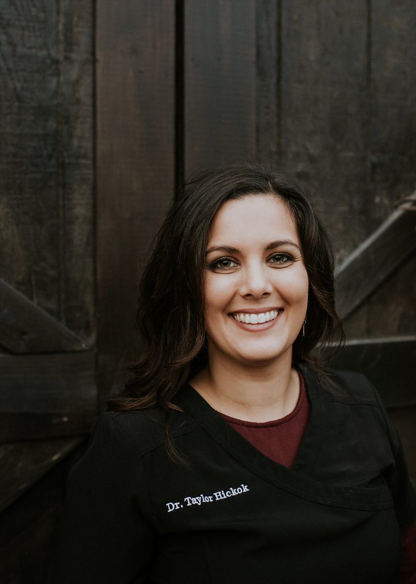 Portrait of Dr. Taylor Hickok, smiling, with dark wavy hair and black scrubs embroidered with her name.