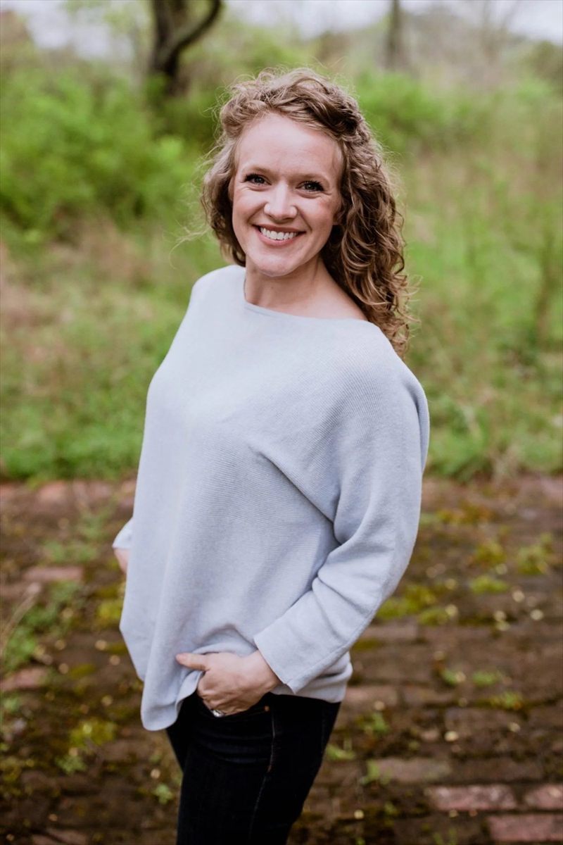 Portrait of Kristin Hunzicker, smiling, with curly reddish-blonde hair, wearing a light blue sweater, outdoors with greenery behind her.